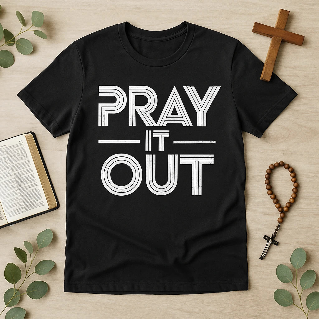 A black t-shirt with the words "PRAY IT OUT" printed in bold, white uppercase letters. The shirt is laid flat on a wooden surface surrounded by a Bible, a wooden cross, a rosary, and some green leaves.
