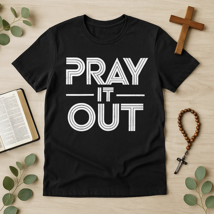 A black t-shirt with the words "PRAY IT OUT" printed in bold, white uppercase letters. The shirt is laid flat on a wooden surface surrounded by a Bible, a wooden cross, a rosary, and some green leaves.