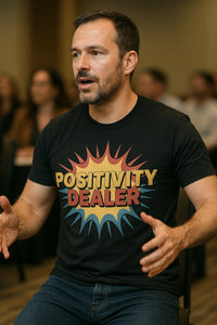 A man with short dark hair and a beard is sitting and gesturing with his hands. He is wearing a black t-shirt with the text 