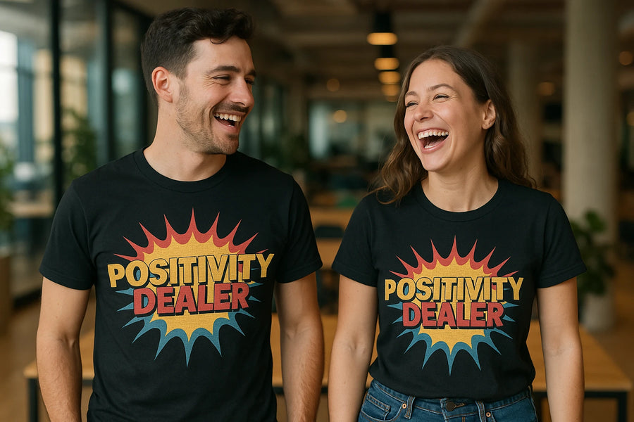 A smiling man and woman stand next to each other, both wearing black t-shirts with the text "POSITIVITY DEALER" in bold, colorful letters surrounded by a burst design. They appear to be in an indoor setting with modern decor and soft lighting.