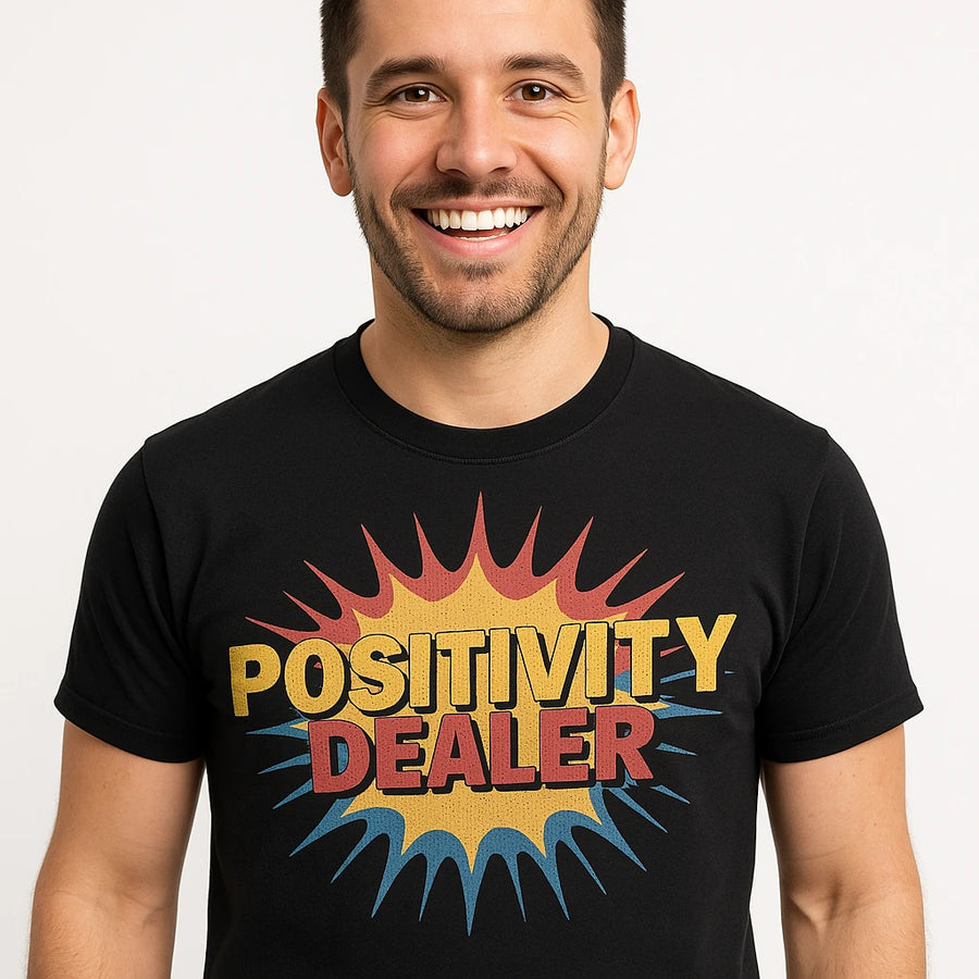 A person with short dark hair and a beard, smiling broadly. They are wearing a black T-shirt with the words "POSITIVITY DEALER" in large, colorful letters over a burst graphic.
