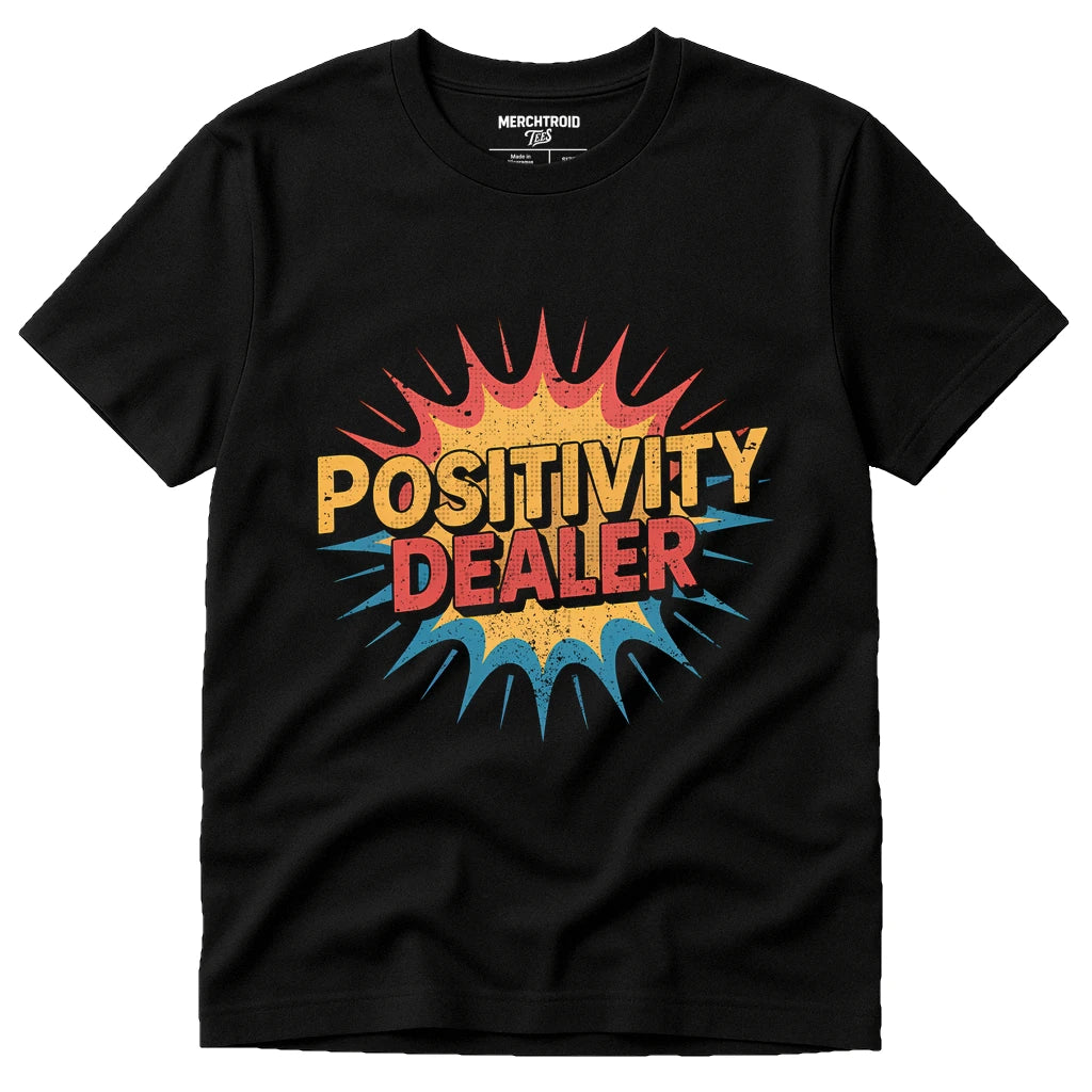 A black t-shirt featuring a colorful, comic-style graphic with the text "POSITIVITY DEALER" in bold letters. The design includes a burst pattern with red, yellow, and blue colors.