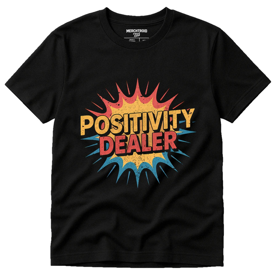A black t-shirt featuring a colorful, comic-style graphic with the text "POSITIVITY DEALER" in bold letters. The design includes a burst pattern with red, yellow, and blue colors.