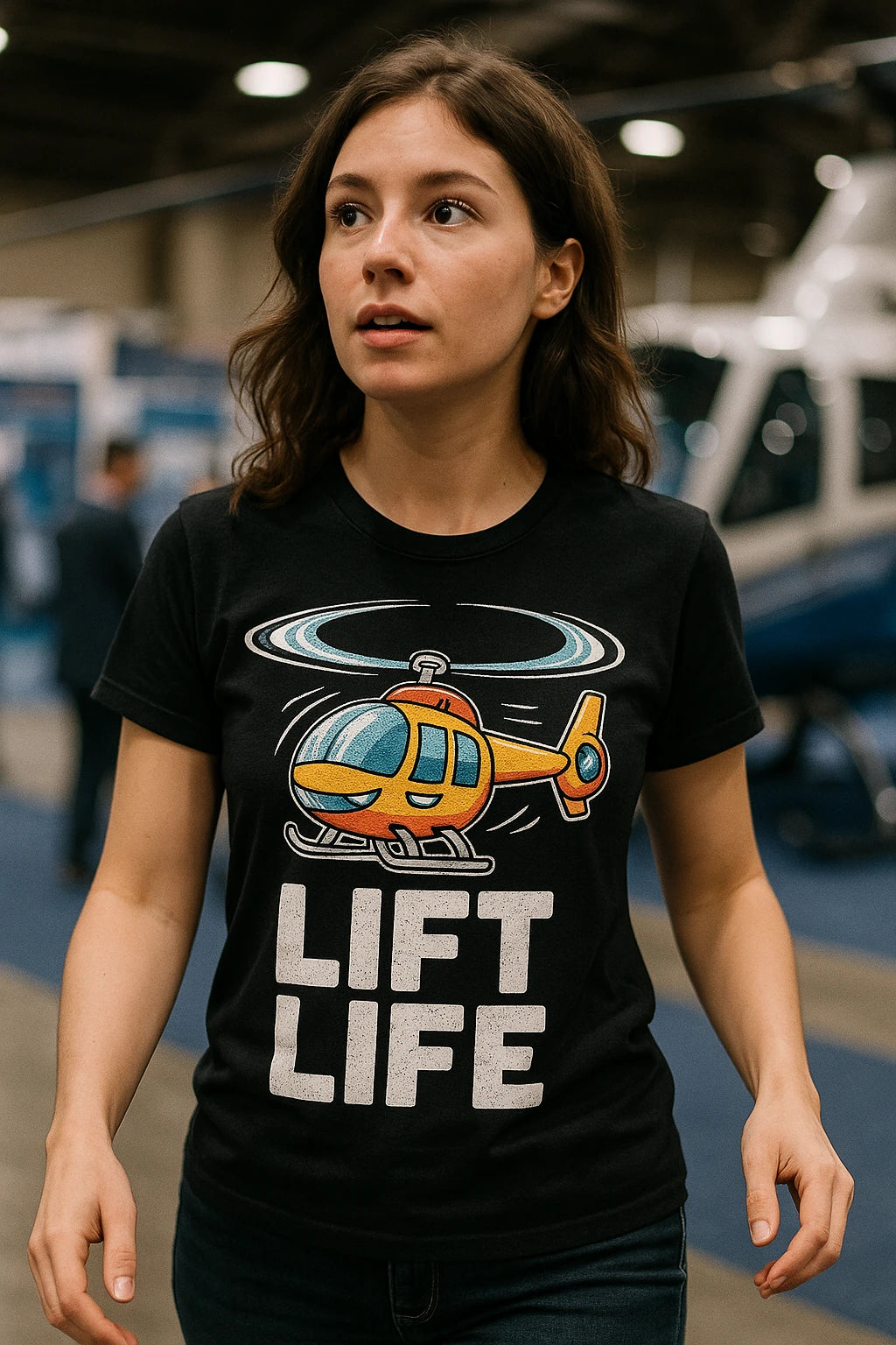 A person with long brown hair is wearing a black T-shirt featuring a colorful cartoon helicopter and the text "LIFT LIFE." The background appears to be an indoor setting with blurred elements suggesting a possible event or exhibition space.