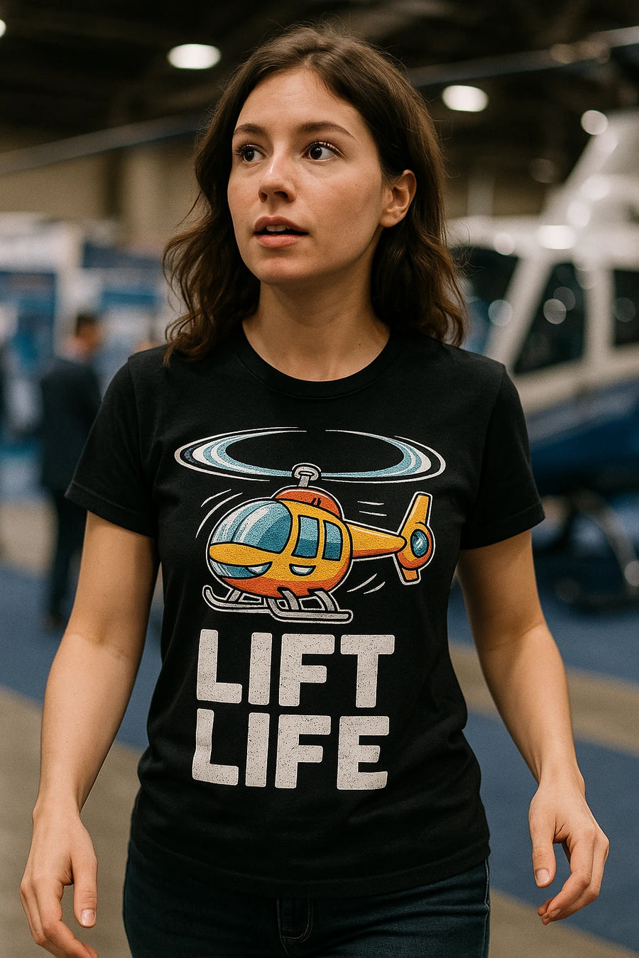 A person with long brown hair is wearing a black T-shirt featuring a colorful cartoon helicopter and the text "LIFT LIFE." The background appears to be an indoor setting with blurred elements suggesting a possible event or exhibition space.