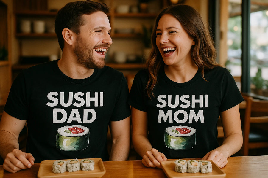 A man and woman are sitting side by side at a table, both wearing black t-shirts with sushi illustrations. The man’s shirt reads “SUSHI DAD” and the woman’s shirt reads “SUSHI MOM.” They are smiling and looking at each other, each with a plate of sushi rolls in front of them.