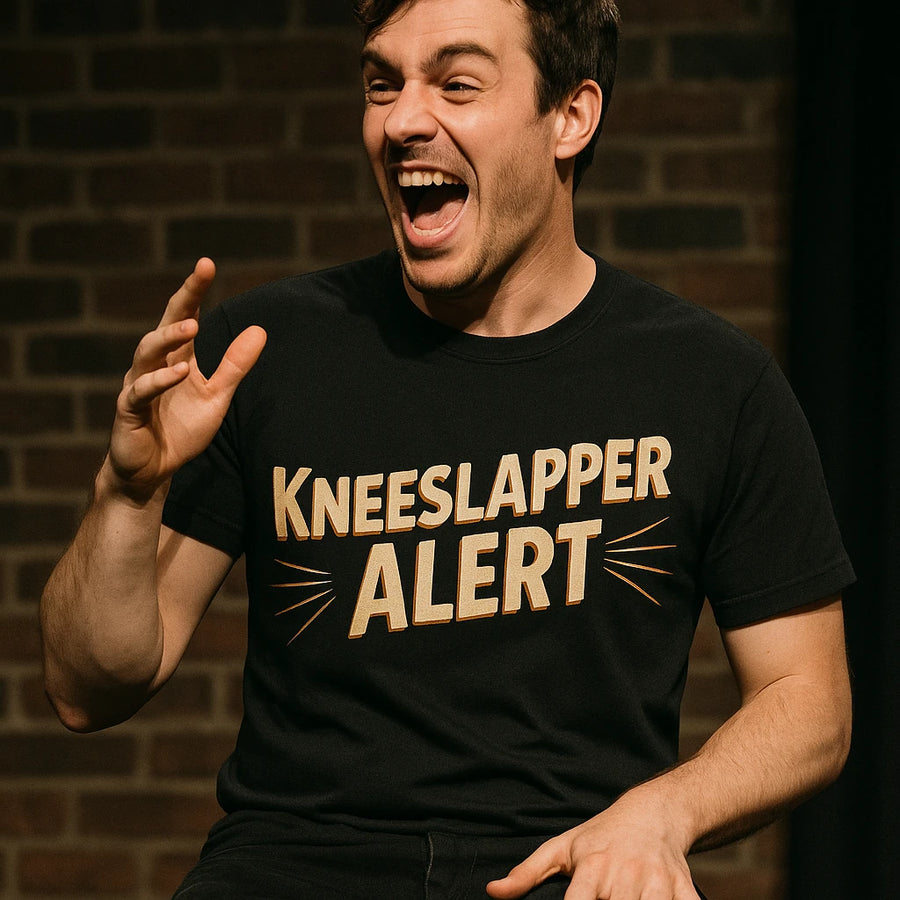 A person with short dark hair is wearing a black t-shirt with the text "KNEESLAPPER ALERT" printed in bold, yellow letters. They are laughing energetically, with one hand raised and the other resting on their knee. The background features a brick wall.