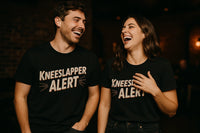 A man and a woman are standing side by side, both laughing joyfully. They are wearing matching black T-shirts with the text 