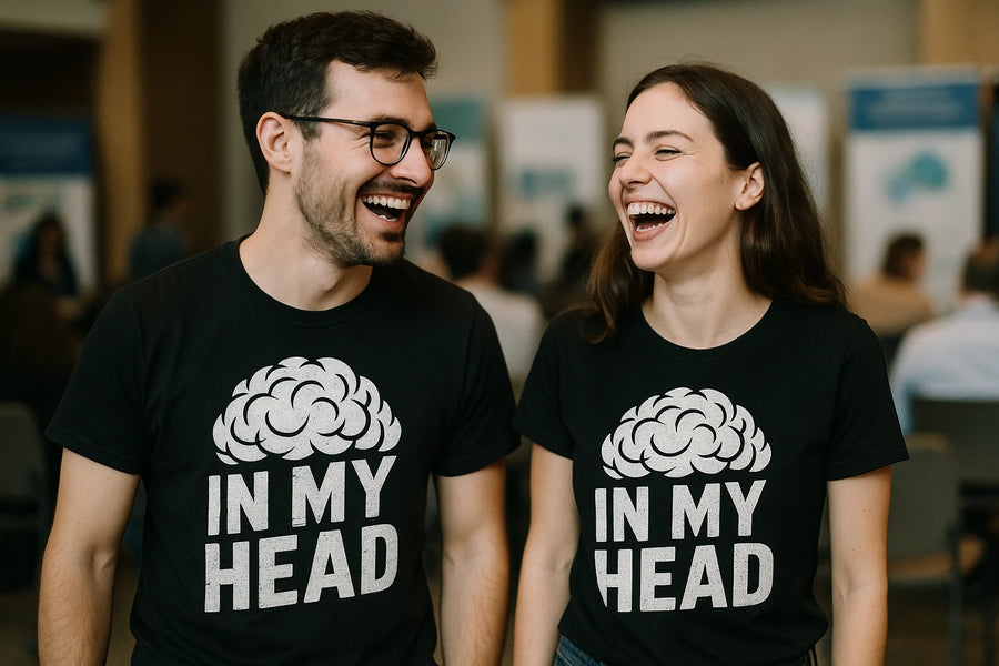 Two people, a man and a woman, are standing indoors and laughing together. Both are wearing black t-shirts with a brain graphic and the words "IN MY HEAD" printed on them. They appear to be at an event or gathering, with a blurred background of people and display boards.
