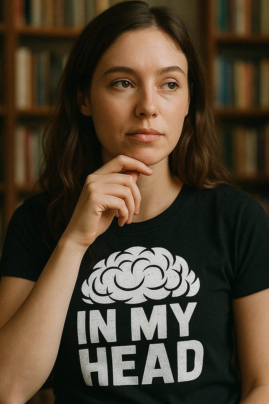 A person with long brown hair is wearing a black T-shirt featuring an illustration of a stylized brain above the words "IN MY HEAD." They are resting their chin on their hand and gazing thoughtfully to the side. In the background, there is a blurred bookshelf.