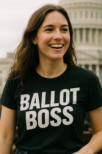 A person with long brown hair is smiling and wearing a black T-shirt with the text 
