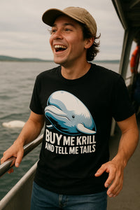 A person with a light brown cap and black T-shirt is smiling widely on a boat deck. The T-shirt features an illustration of a whale with the text 