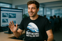 A person with short brown hair and a beard is sitting at a desk in an office environment, smiling and gesturing with one hand. They are wearing a black t-shirt featuring a cartoonish beluga whale and text that reads 
