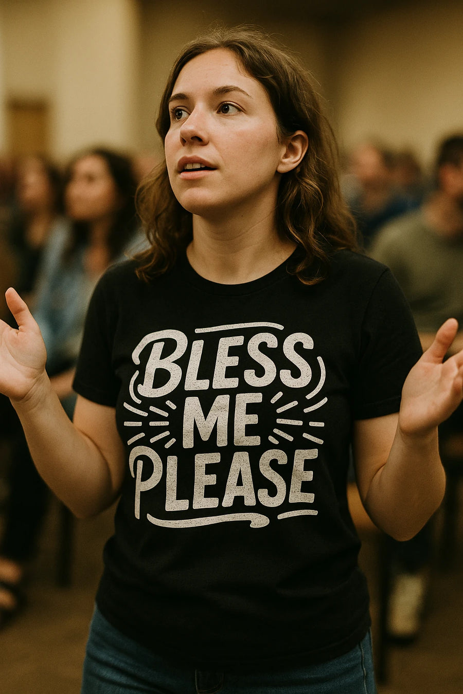 A person with wavy brown hair is standing with their hands raised in a gesture similar to prayer or worship. They are wearing a black T-shirt with the text "BLESS ME PLEASE" printed on it. The background is blurred, showing other people in a room.