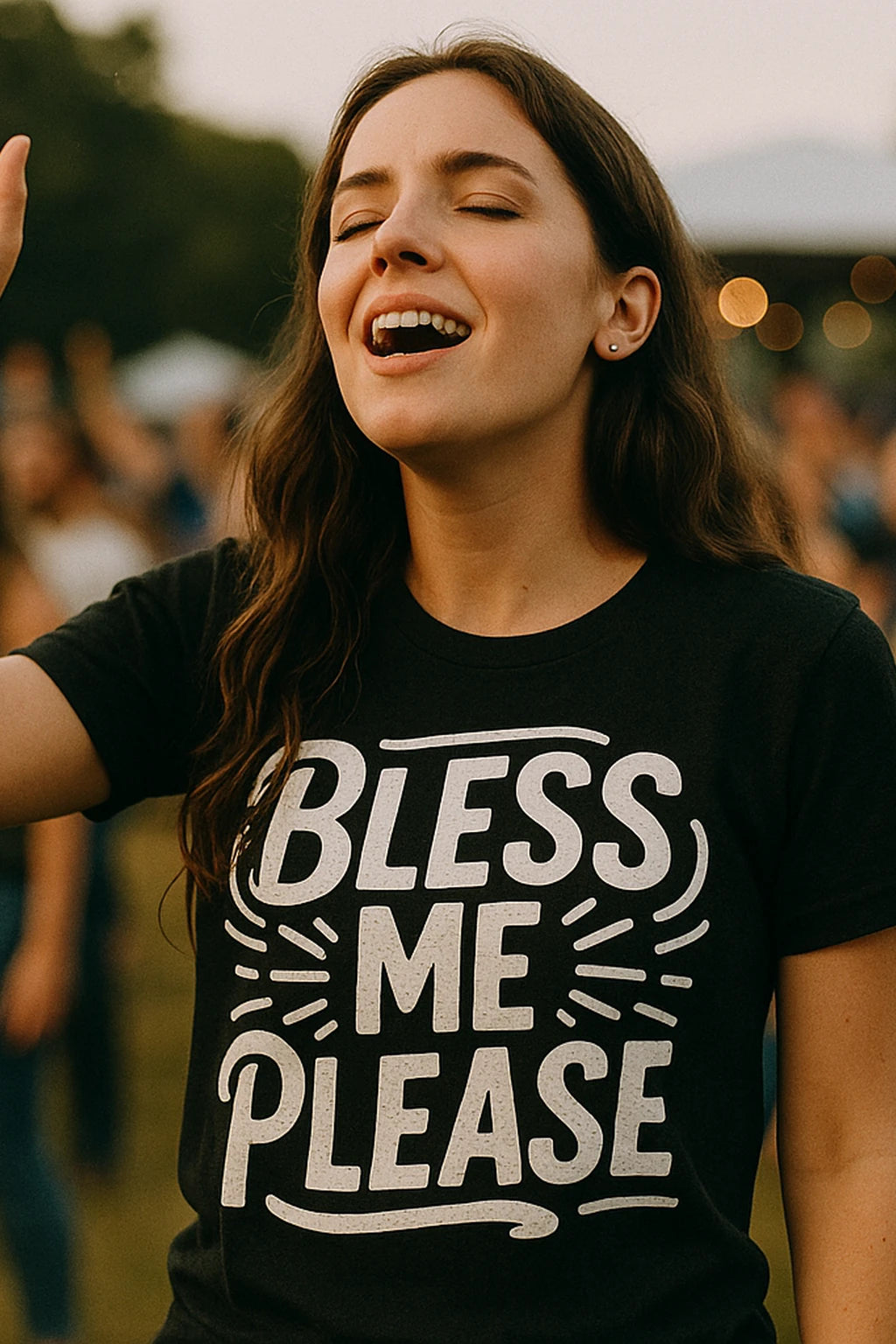 A person with long brown hair is wearing a black T-shirt with the text "BLESS ME PLEASE" in bold white letters. They have their eyes closed and appear to be singing or speaking with expression. The background is slightly blurred, suggesting an outdoor event or gathering.