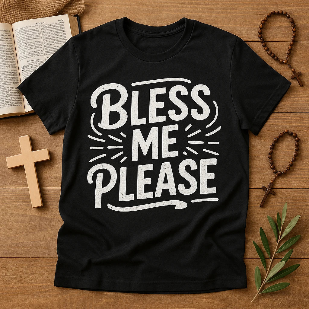 A black t-shirt with the text "BLESS ME PLEASE" in bold white letters, surrounded by decorative lines. The shirt is laid flat on a wooden surface, accompanied by an open book, a wooden cross, two rosaries, and a sprig of green leaves.