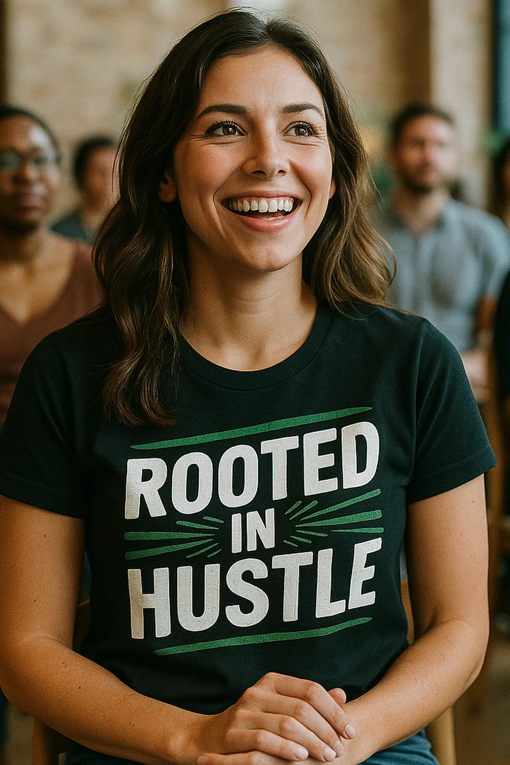A person with long brown hair is smiling and wearing a black T-shirt that reads "ROOTED IN HUSTLE" in bold, white and green lettering. The background shows other people slightly out of focus, suggesting a group setting.