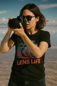 A person wearing sunglasses, holding a camera up to their eye, is outdoors. They are wearing a black t-shirt with a graphic of a camera and the words 
