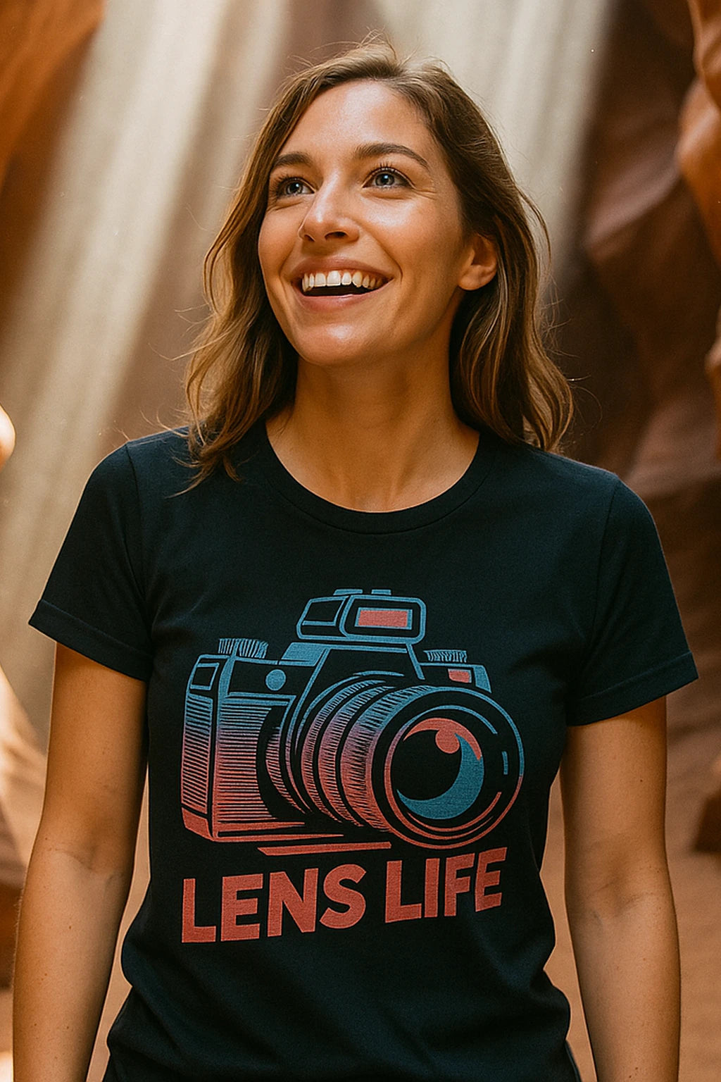 A person with long hair and a cheerful expression is wearing a black T-shirt featuring a colorful camera graphic and the words "LENS LIFE." They are standing in a naturally lit environment.
