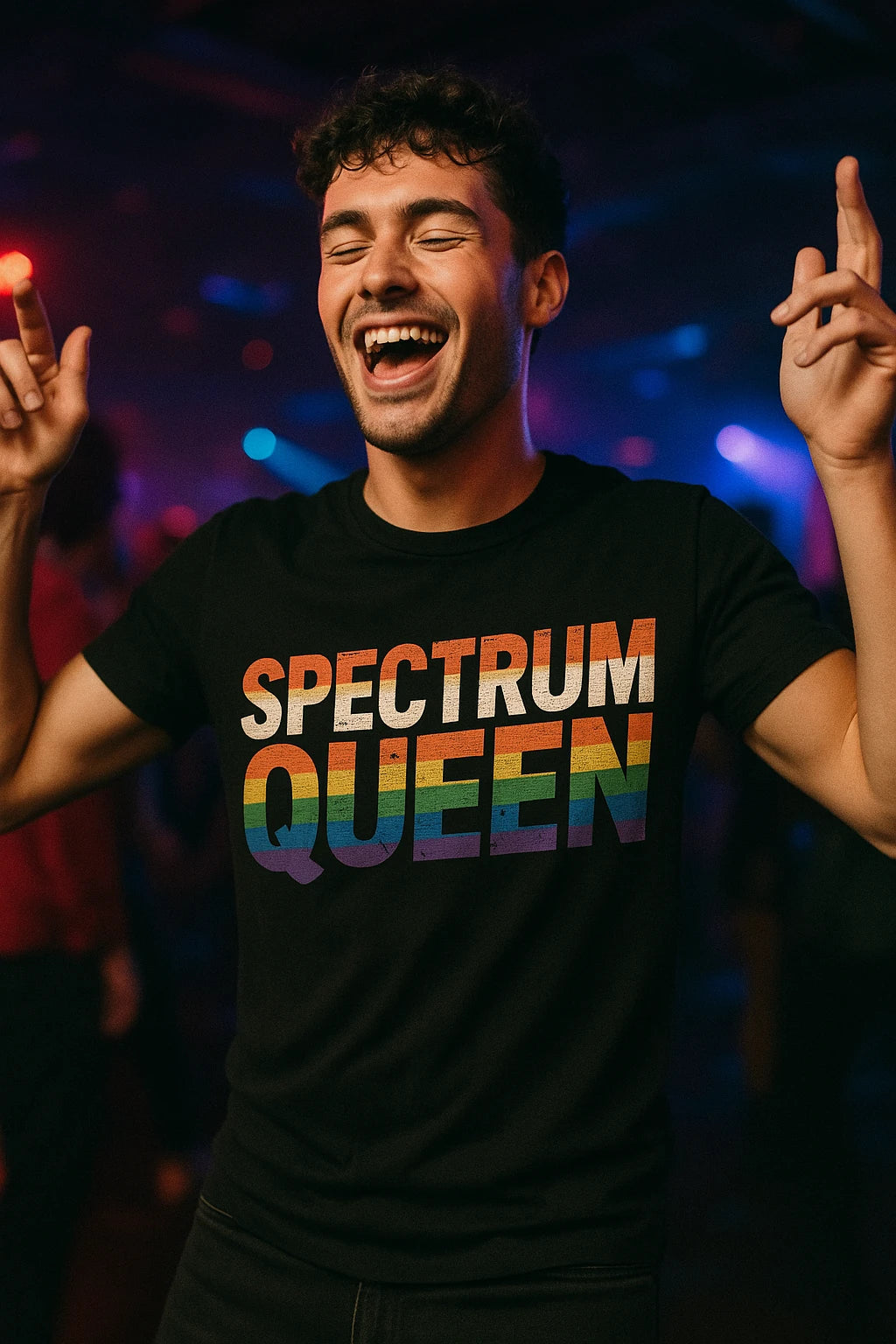 A person wearing a black t-shirt with the text "SPECTRUM QUEEN" in rainbow colors. They are smiling broadly and holding up both hands with fingers pointing upwards. The background is a lively, colorful setting, suggesting a party or nightclub.