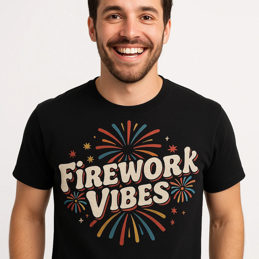 A person with short dark hair and a beard is smiling widely. They are wearing a black t-shirt with colorful fireworks graphics and the words "Firework Vibes" printed on it. The background is a plain, light color.