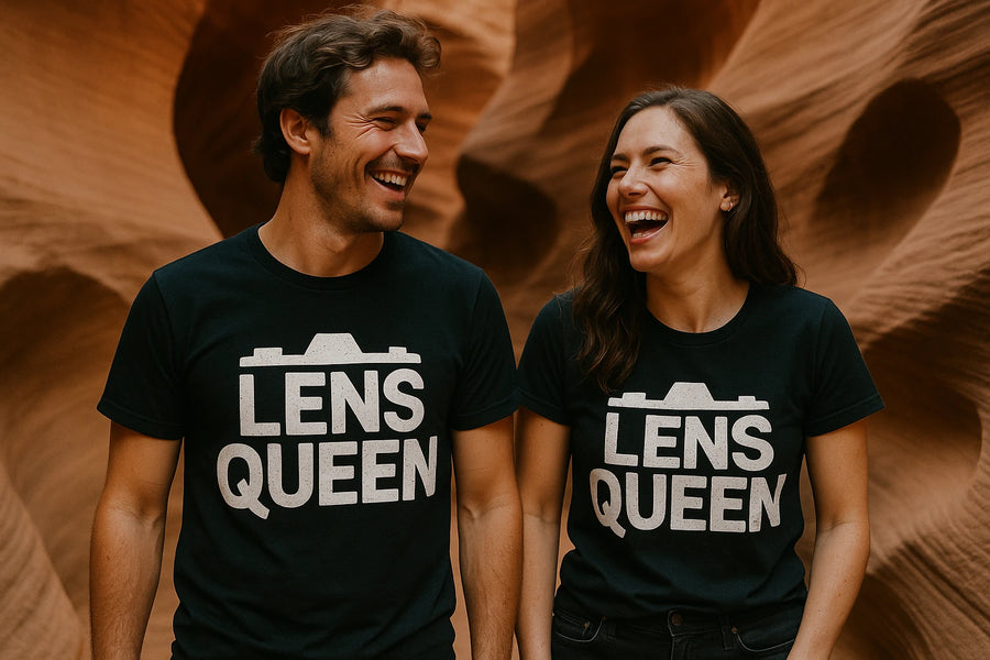 A man and a woman are standing side by side, both wearing black T-shirts with the words "LENS QUEEN" printed on them in white with a camera silhouette above the text. They are smiling and looking at each other in front of a textured, sandstone wall.