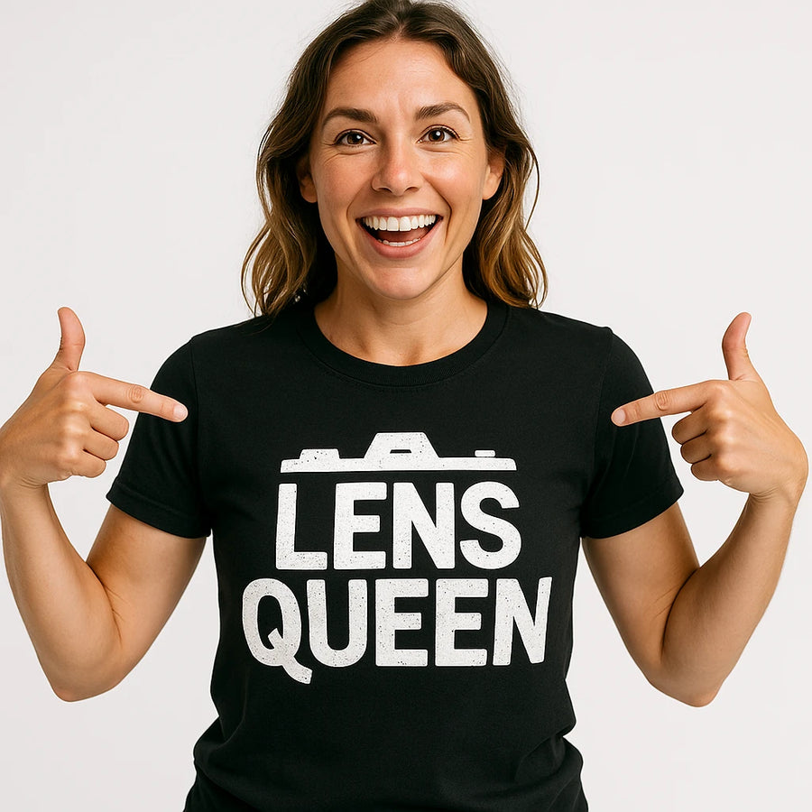A person with long brown hair smiling and pointing at their black t-shirt, which has the text "LENS QUEEN" printed in white.