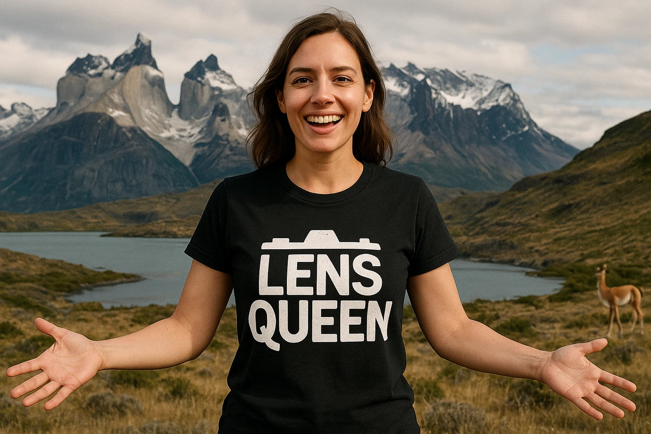 A person with dark hair and wearing a black t-shirt that reads "LENS QUEEN" stands smiling with arms outstretched. In the background, there is a scenic view of mountains, a lake, and a grassy landscape, with a guanaco visible on the right.