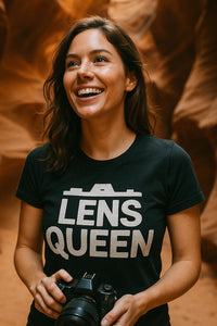 A woman with long brown hair is smiling while holding a camera. She is wearing a black t-shirt with the text 
