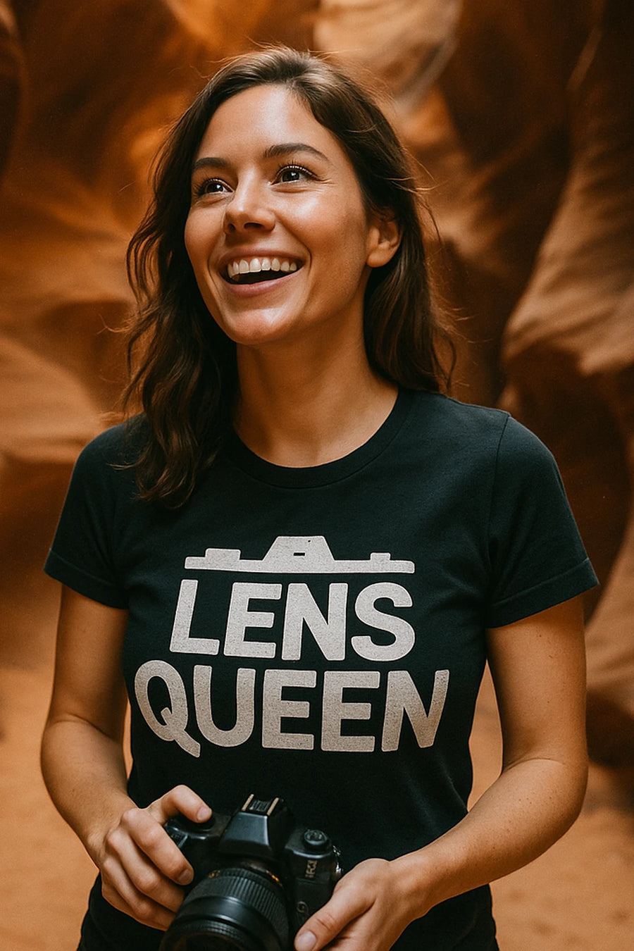 A woman with long brown hair is smiling while holding a camera. She is wearing a black t-shirt with the text "LENS QUEEN" printed on it. The background features a warm-toned rock formation, suggesting a natural or canyon setting.