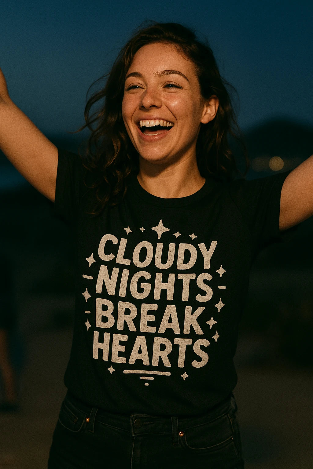 A person with long hair is smiling widely with their arms raised. They are wearing a black t-shirt with white text reading "CLOUDY NIGHTS BREAK HEARTS" surrounded by small star designs. The background is dimly lit, suggesting it's nighttime.