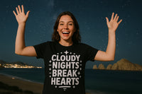 A person smiling brightly under a starry night sky, raising their arms joyfully. They are wearing a black t-shirt with the text 