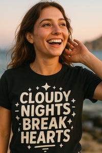 A person with long brown hair smiles broadly while touching their hair. They are wearing a black T-shirt with the text 