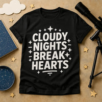 A black t-shirt lies on a tan surface. The shirt features bold white text and star graphics. Surrounding items include a small telescope, constellation books, star cutouts, and a fluffy cloud cutout, creating an astronomy-themed setting.