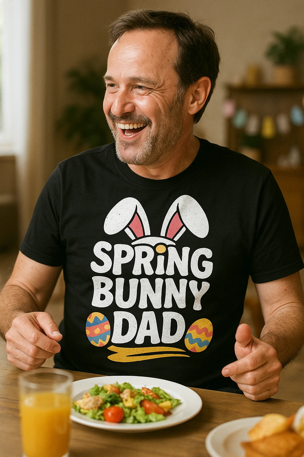 A man with short hair and a beard is sitting at a table, smiling and gesturing with his hands. He is wearing a black t-shirt with a design that includes bunny ears and colorful Easter eggs. On the table in front of him are a plate of salad and a glass of orange juice. The background shows a bright room with some Easter decorations.