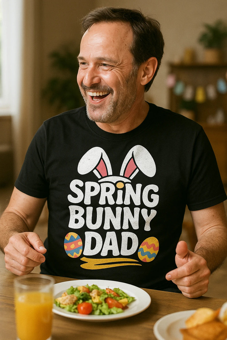 A man with short hair and a beard is sitting at a table, smiling and gesturing with his hands. He is wearing a black t-shirt with a design that includes bunny ears and colorful Easter eggs. On the table in front of him are a plate of salad and a glass of orange juice. The background shows a bright room with some Easter decorations.
