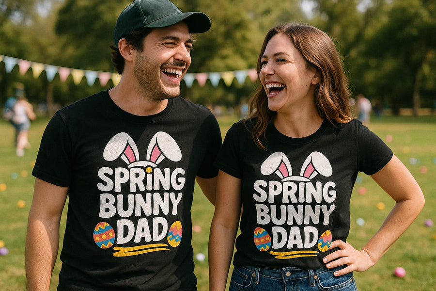 A man and woman are standing outdoors on a sunny day, laughing and looking at each other. They are both wearing black T-shirts with the text "SPRING BUNNY DAD" along with images of bunny ears and colorful Easter eggs. The setting appears to be a park with a blurred background of grass, trees, and colorful bunting flags. The man is wearing a dark cap.