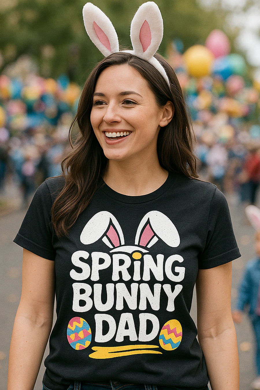 A person smiling and wearing a black t-shirt with "SPRING BUNNY DAD" written on it, alongside bunny ears and decorated eggs. The person is also wearing white bunny ear headbands. The background is blurred with colorful balloons and people.