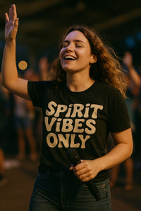 A person with wavy hair is smiling and holding a microphone. They are wearing a dark T-shirt with the words 