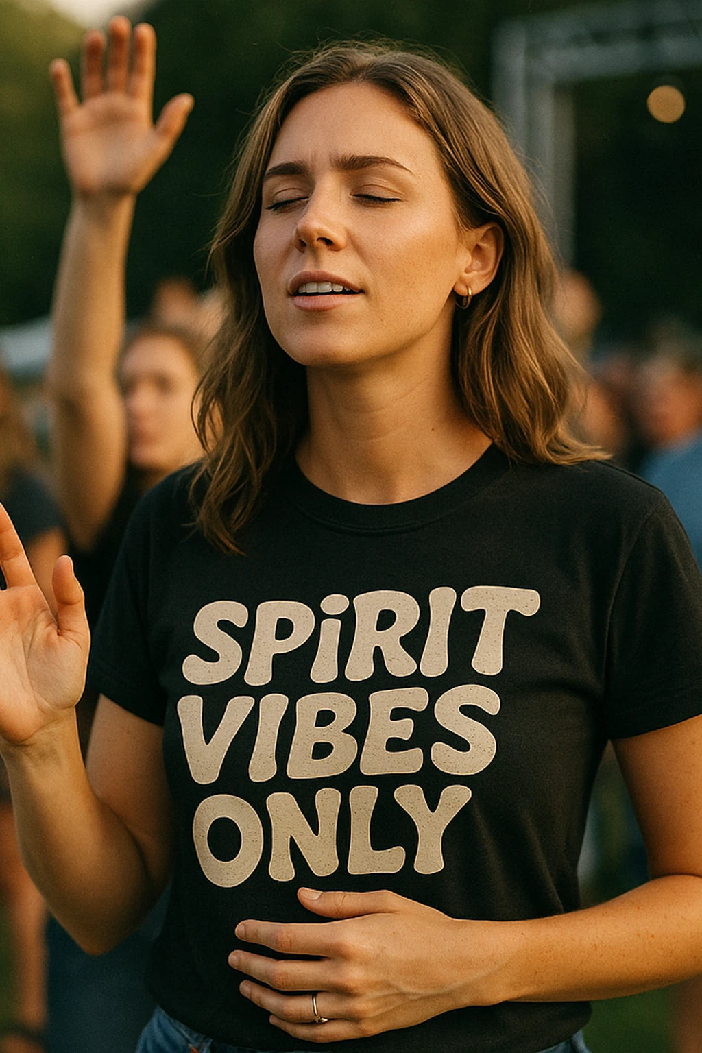 A person with long brown hair is wearing a black t-shirt with the text “SPIRIT VIBES ONLY.” The person appears to be outdoors, with eyes closed and a serene expression, possibly participating in an event with a crowd in the background.