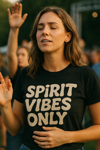 A person with long brown hair is wearing a black t-shirt with the text “SPIRIT VIBES ONLY.” The person appears to be outdoors, with eyes closed and a serene expression, possibly participating in an event with a crowd in the background.