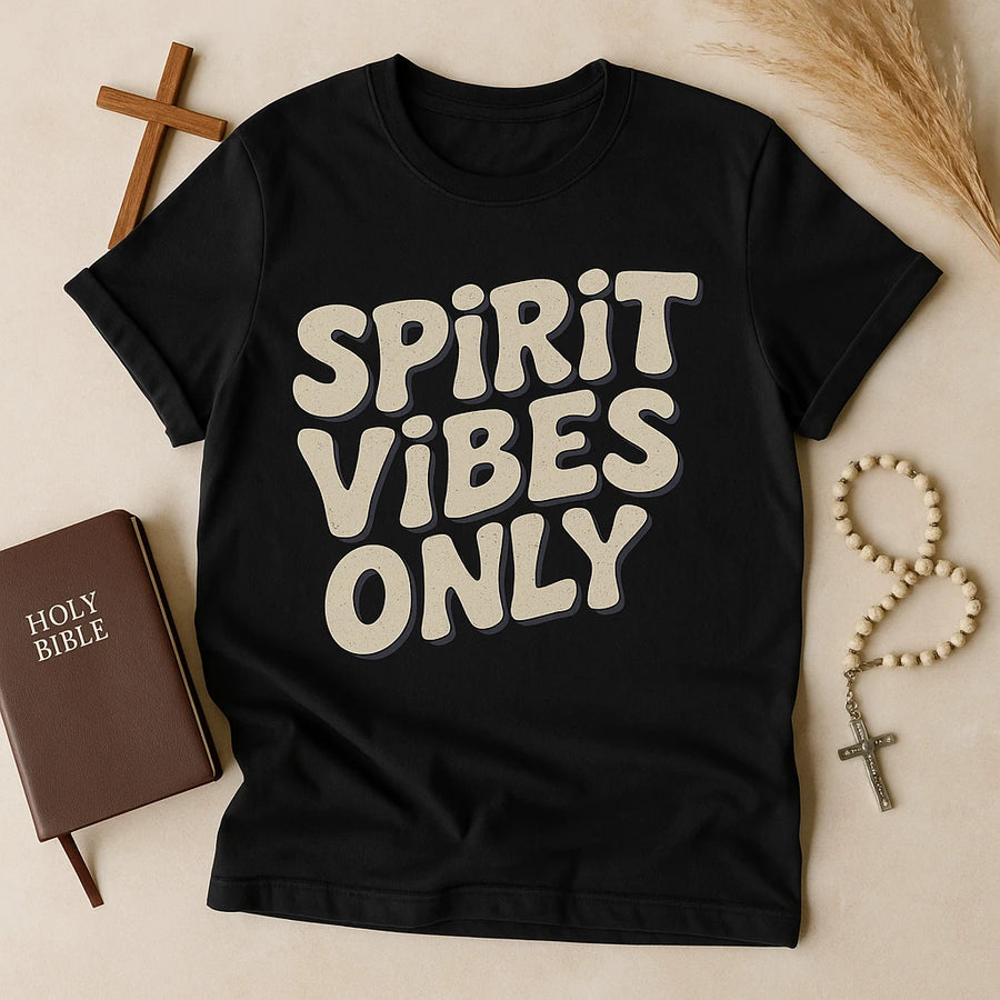 A black t-shirt with the text "SPIRIT VIBES ONLY" in bold, light-colored font. It is laid flat on a surface next to a brown Holy Bible, a wooden cross, and a string of rosary beads. Dried plants are visible in the background.