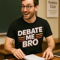 A person with glasses, a beard, and short hair is sitting at a desk. They are wearing a black T-shirt with the text 