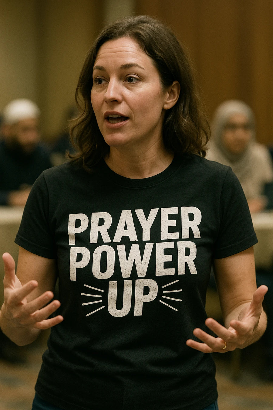 A person with shoulder-length brown hair is wearing a black T-shirt with the words "PRAYER POWER UP" printed on it. They appear to be speaking, with their hands gesturing. In the background, there are other people sitting in a room, some wearing headscarves.