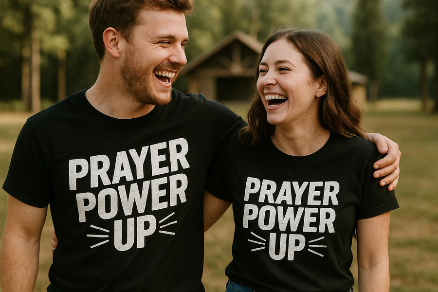 A man and a woman are standing side by side, both laughing and wearing black t-shirts that read "PRAYER POWER UP." They are outdoors, with greenery and a wooden shelter visible in the background. The woman has her arm around the man's shoulder, and they appear to be enjoying each other's company.