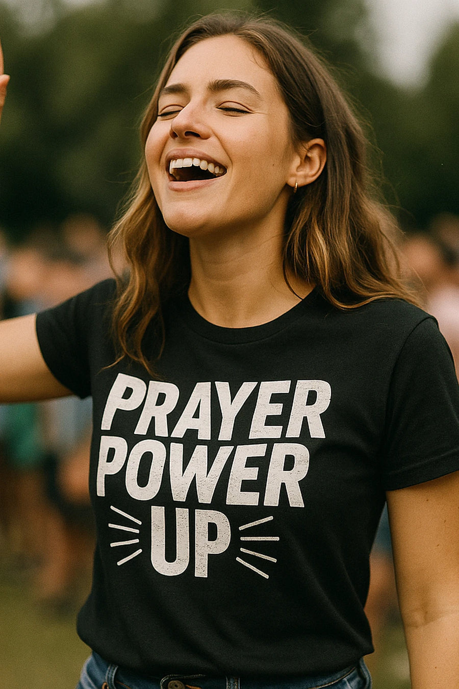 A person with long hair is smiling widely outdoors, wearing a black T-shirt with the text "PRAYER POWER UP" printed on it in bold white letters. The background is blurred with people and greenery.