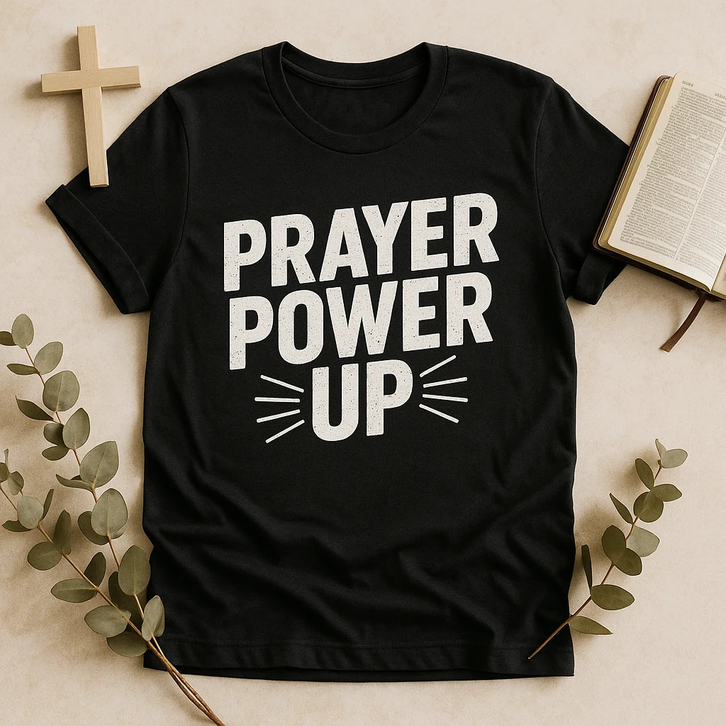 A black T-shirt with the words "PRAYER POWER UP" in bold, white, capital letters. The shirt is placed on a flat surface alongside a wooden cross, an open book, and some green foliage branches.