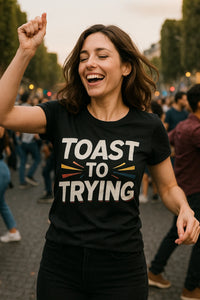 A person with shoulder-length brown hair is smiling broadly and raising one arm, appearing joyful. They are wearing a black T-shirt with the text 