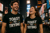 A man and a woman are standing in a lively urban setting, possibly a city center at night, laughing together. Both are wearing black T-shirts with the phrase 