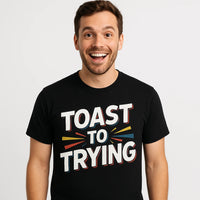 A person with short brown hair and a beard is smiling widely. They are wearing a black T-shirt with the text 